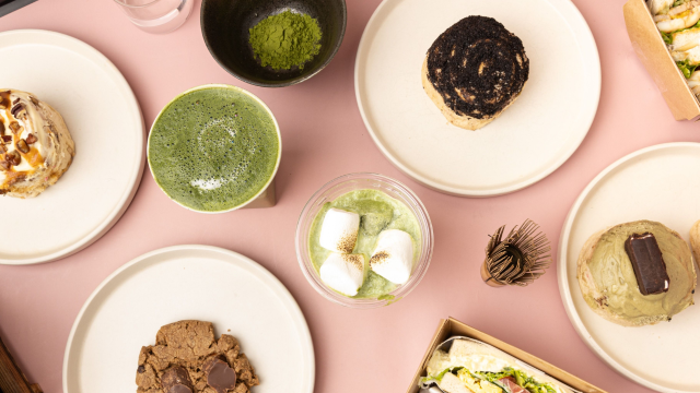 A pink table with different pastries and matcha drinks on it. There is also a matcha whisk and a bowl of matcha powder displayed.